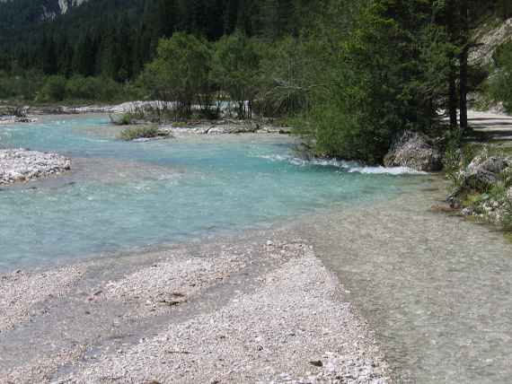Das Bild zeigt einen Fluss mit Kiesbänken, der weitgehend frei fließen kann. Nur rechts ist eine Straße, die mit einigen großen Steinen geschützt ist.