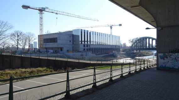 Schräger Blick zum Neubau, aufgenommen unterhalb der Autobahn, davor verläuft der Donaukanal. Zwei Baukräne sind jeweils vor und hinter dem Neubau zu sehen. Die Außenfassade nimmt Form an. Ganz rechts ist die Eisenbahnbrücke zu sehen bei sonnigem und trockenen Wetter.