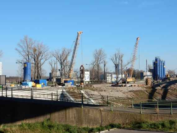 Vier hohe Betonbehälter, zwei Kräne und einige Baucontainer können auf dem Foto entnommen werden.