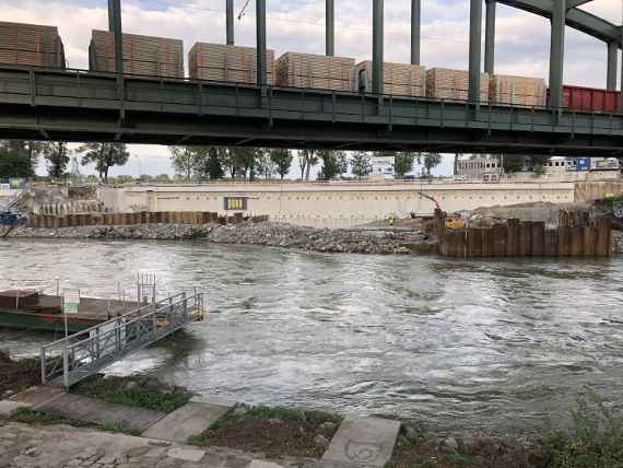 Der Donaukanal verläuft von links nach rechts, darüber verläuft eine Eisenbahnbrücke, wo gerade ein Güterzug durchfährt. Auf der anderen Seite des Donaukanals ist eine Betonwand zu erkennen, davor in den Donaukanal eingerammte Spundwände.