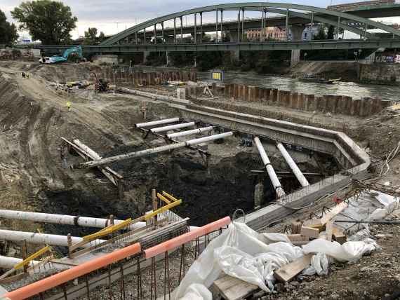 Man sieht die Baugrube, zwei Wände in Beton und Stahlrohre zur Abstützung der Baugrube. Rechts verläuft der Donaukanal und darüber die Eisenbahnbrücke.