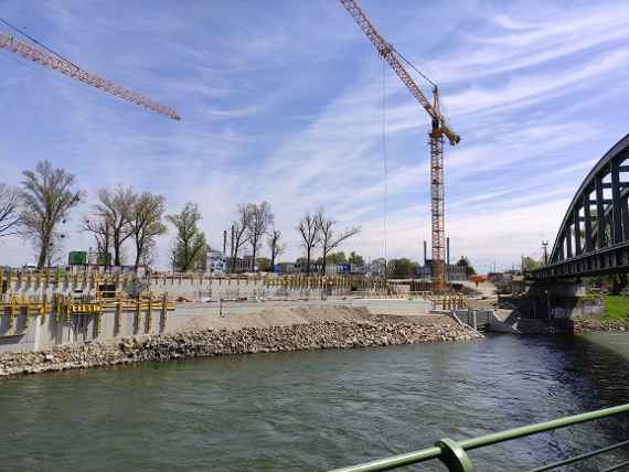 Blick über Donaukanal. Zwei Kräne stehen links und rechts auf der Baustelle, ganz rechts die Eisenbahnbrücke. Etwas wolkig, sonniges und trockenes Wetter.