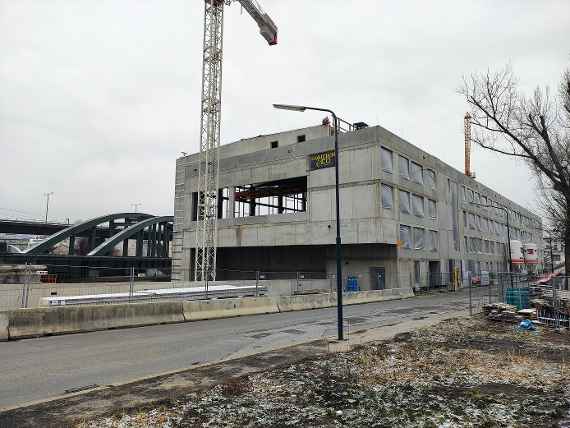 Blick zum Neubau. Rechts neben Neubau verläuft eine Straße. Alle Fensteröffnungen sind provisorisch mit Planen zugedeckt (Schutz vor Kälte). Vor dem Bau steht ein Kran, links ist die Eisenbahnbrücke zu sehen. Wolkiges und kaltes Wetter knapp über dem Gefrierpunkt.