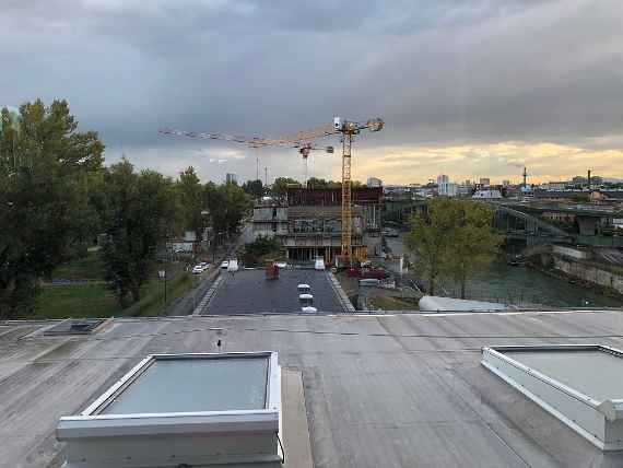 Man sieht die Baustelle mit 2 großen Baukränen, links die Straße und rechts den Donaukanal. Im Hintergrund die Häuser von Wien. Wolkiges Wetter, aber trocken.