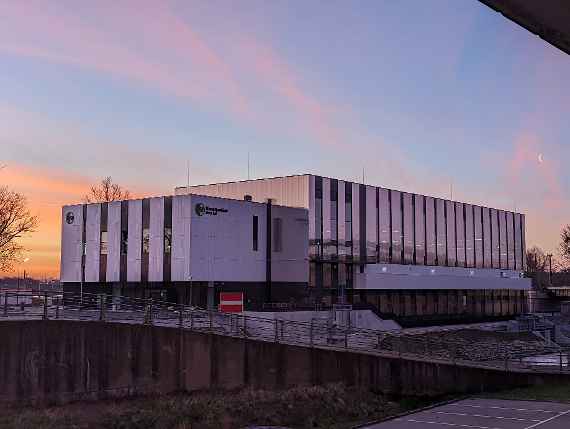 Schräger Blick zum Neubau über den Donaukanal, aufgenommen unterhalb der Autobahn. Es sind das Logo „BOKU“ sowie die Beschriftungen „Wasserbaulabor“ und „River Lab“ zu lesen. Bildaufnahme erfolgte früh morgens beim Sonnenaufgang, daher gelb-rötlich-blauer Himmel mit etwas Wolken. Schaut man aber ganz rechts auf dem Foto, sieht man deutlich der abnehmenden Mondsichel etwas mit Wolke verdeckt. Oben rechts sieht man einen kleinen Teil der Autobahn. Da das Gebäude seitlich am Donaukanal spiegelt, sieht man darauf ein Teil der Eisenbahnbrücke.