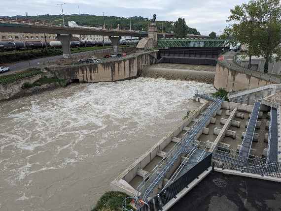 Das Bild zeigt die Wehranlage frontseitig, rechts der Fischaufstieg. Die Aufnahme erfolgte am 30. August 2023, nachdem in den letzten Tagen vorher im Westen Österreichs durch Unwetter, die aus Italien kamen Hochwasseralarm herrschte. Man sieht sehr trübes Wasser auf dem Donaukanal. Zwei Hubschwenktore sind abgesenkt, um den Durchfluss zu begrenzen und das Treibgut, im Wesentlichen Holz von der Wasseroberfläche zu sammeln. Links die Autobahnbrücke und dahinter der Bahnhof Nußdorf.