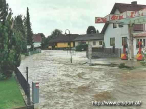 Stark strömender Fluss durch den Ort Haitzendorf, am linken Bildrand ist ein Garten mit Bäumen sichtbar, rechts einige Häuser, und dazwischen fließt ein stark gewellter schmutzig brauner Fluss, wo normalerweise die Straße entlang führt.