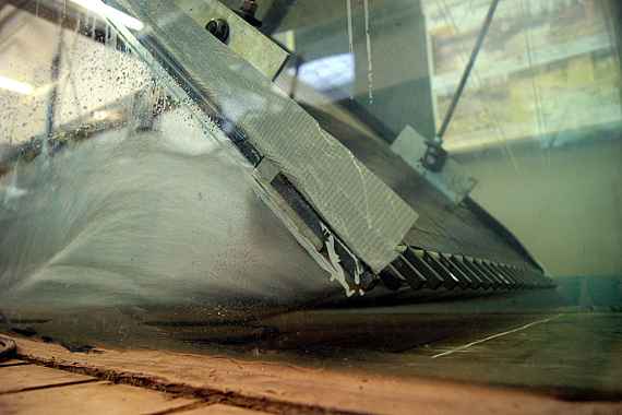 Durch eine Glaswand sieht man, wie an einer Wand aufgestautes Wasser durch einen Spalt am Boden mit hoher Geschwindigkeit abfließt.