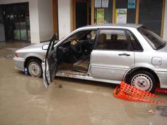 Ein Auto mit offener Fahrertür unter dem Hochwasser, dahinter zwei undefinierte Einkaufsgeschäfte.