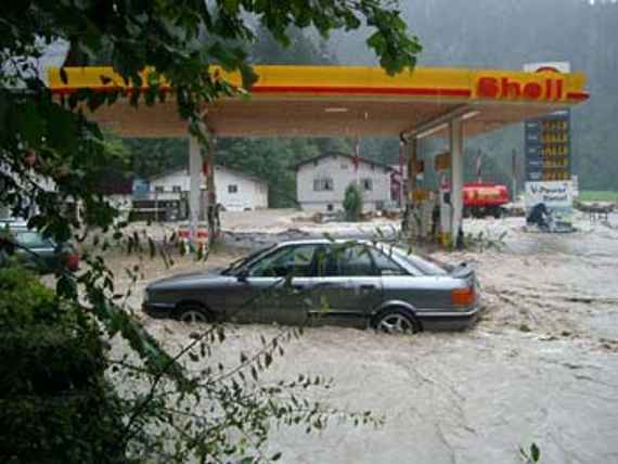 Eine überflutete Shell-Tankstelle, dahinter zwei Häuser. Reißendes Wasser und ganz links ein Baum.