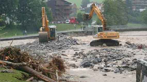 Bregenzerach im Abschnitt Reuthe-Bezau mit Ufersicherung, 2 Bagger stehen auf Schotter, ringsum fließt das derzeit schmutzige Wasser. Links sind etwas Anlandungen zu sehen. Dahinter einige Bäume und zwei Häuser.