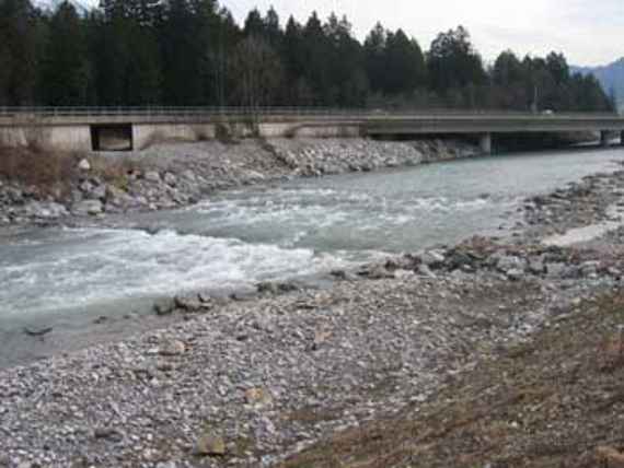 Fluss ILL im Bereich von Nenzing flussab der Frutzmündung beim Blick gegen Fließrichtung. Vorne Ufer, hinten Ufer mit Strecke und Brücke, dahinter Waldgebiet.