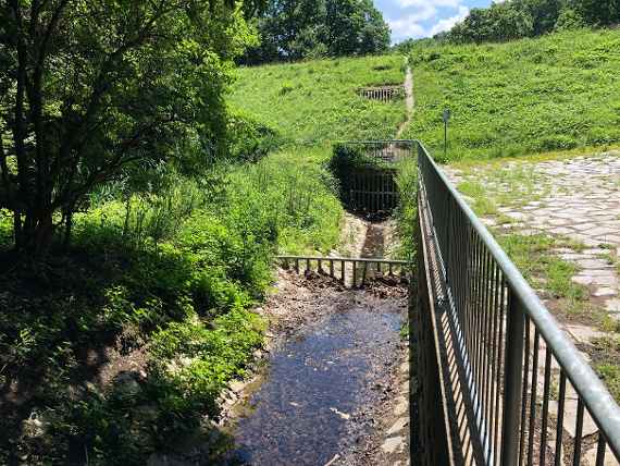 Man sieht einen mit Gras bewachsenen Damm und einen kleinen Bach der zu diesem Damm fließt. Unten ist im Damm eine Öffnung für den Grundablass. Weiter oben eine zweite Öffnung für die Hochwasserentlastung mit dem Heber.