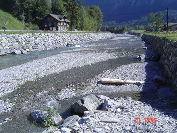 Fluss mit Anlandungen, links ist ein Haus. Fotoaufnahme bei sonnigem Wetter.