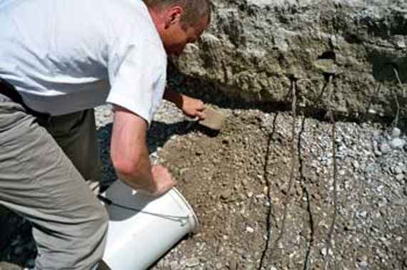 Entnahme von Unterschichtmaterial aus einem Renaturierungsabschnitt an der Isar. Diplom-Ingenieur Schaipp schiebt mit einer Schaufel Kies von einem Flussufer in einen Kübel. Die Kiesschicht ist unterteilt – im oberen Teil ist feineres und verfestigtes Material, durchsetzt mit Pflanzenwurzeln sichtbar, im unteren Teil hingegen gröberer, lockerer Kies.