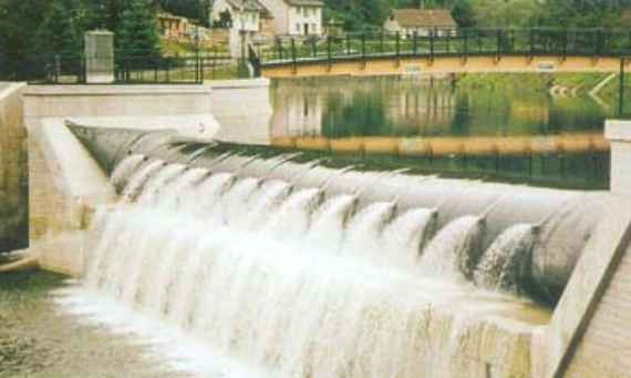 Wasser strömt über die Wehranlage, im Hintergrund sind eine Brücke und dahinter etwas seitlicher 4 Einzelhäuser zu sehen.