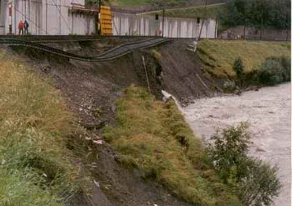 Beschädigte Eisenbahnstrecke der österreichischen Bundesbahn entlang der Salzach nach Hochwasser 1985, der rechte Bildrand zeigt einen Teil der hochwasserführenden Salzach. Das Flusstal ist an dieser Stelle sehr eng. Durch das Hochwasser kam es zur Ufererosion, große Stück des grasbewachsenen Ufers sind in den Fluss gerutscht, der Schienenstrang der Eisenbahnlinie ist unterspült und hängt in der Luft.