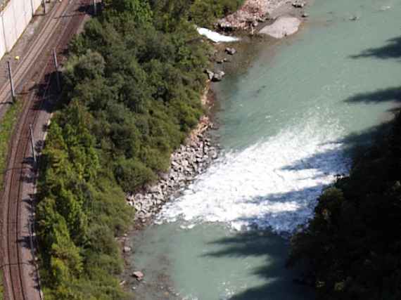 Das Foto zeigt eine der im Modellversuch entwickelten Rampen, die die Flusssohle der Salzach stabilisieren und damit die parallellaufende Eisenbahnstrecke vor Erosion schützen. Man sieht die Eisenbahnstrecke. Über die Rampe fließt das Wasser schneller und ist durch die eingemischte Luft weiß gefärbt.