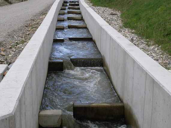 Der Beckenpass besteht aus zwei parallel verlaufenden Betonmauern, die im Grundriss zuerst eine leicht Kurve machen, dann folgt eine 180 Grad Kurve. Durch Querriegel aus Reihen großer Steine entstehen mit Wasser gefüllte Becken. Von Becken zu Becken fließt das Wasser stufenförmig nach unten. Links sieht man eine Böschung mit Wiese und weiter hinten einige Bäume und mehrere Häuser. Hinter dem Fischaufstieg steht ein großes Gebäude (das Kraftwerk).