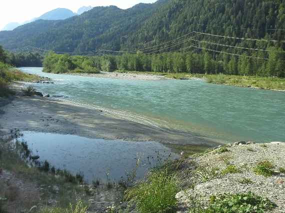 Das Bild zeigt einen Fluss, der von rechts nach links fließt. Entlang des rechten Ufers verläuft eine Stromleitung. Dahinter ist ein mit Wald bewachsener Berg. Am linken Flussufer sieht man eine große Fläche mit feinem Sand und davor eine Fläche mit grobem Kies.