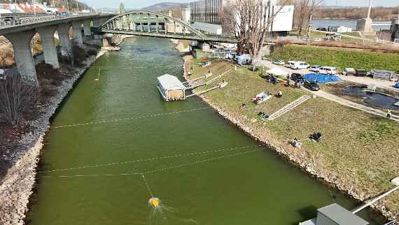 Das Bild zeigt eine Drohnenaufnahme des Donaukanals. Über den Fluss sind drei Seile gespannt. Einige Personen halten sich am Ufer auf. Über das Wasser bewegen sich zwei ADCPs.