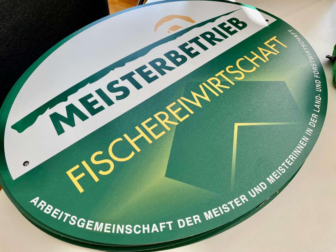 The photo shows a logo taken at an angle, which is circular. In the upper half of the circle, on a white background, the German text "Meisterbetrieb" (English for „master craftsman's business“) is written in green letters. Above the text is a green line and above it a small sun. The lower half of the circle has a green background with the yellow German text " Fischereiwirtschaft" (English for “fishery industrie”). Along the lower half of the circumference is the white German text "Arbeitsgemeinschaft der Meister und Meisterinnen in der Land- und Forstwirtschaft" (English for „Association of master craftsmen and women in agriculture and forestry“). All texts are written in capital letters.