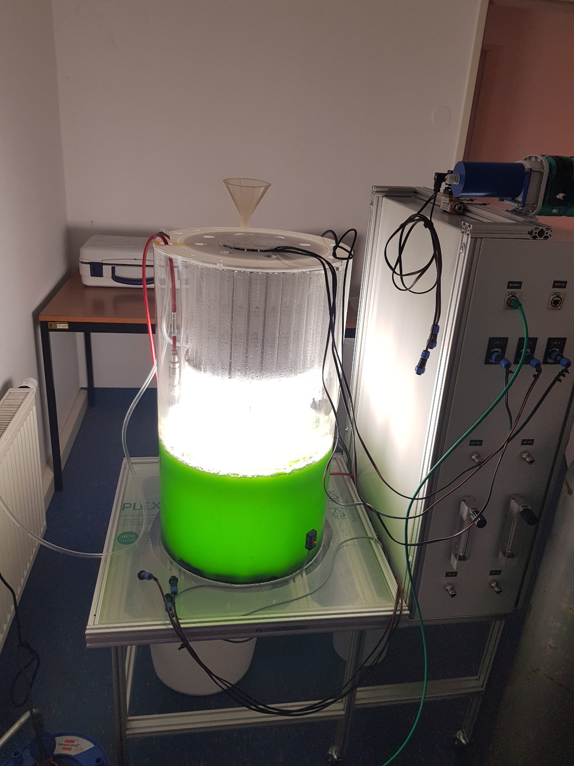 On photo you can see a photobioreactor. In a glass container there is a green liquid at the bottom, namely that of the algae. Above is an illuminator, which is directed to this liquid. Next to it on the right is a technical device.