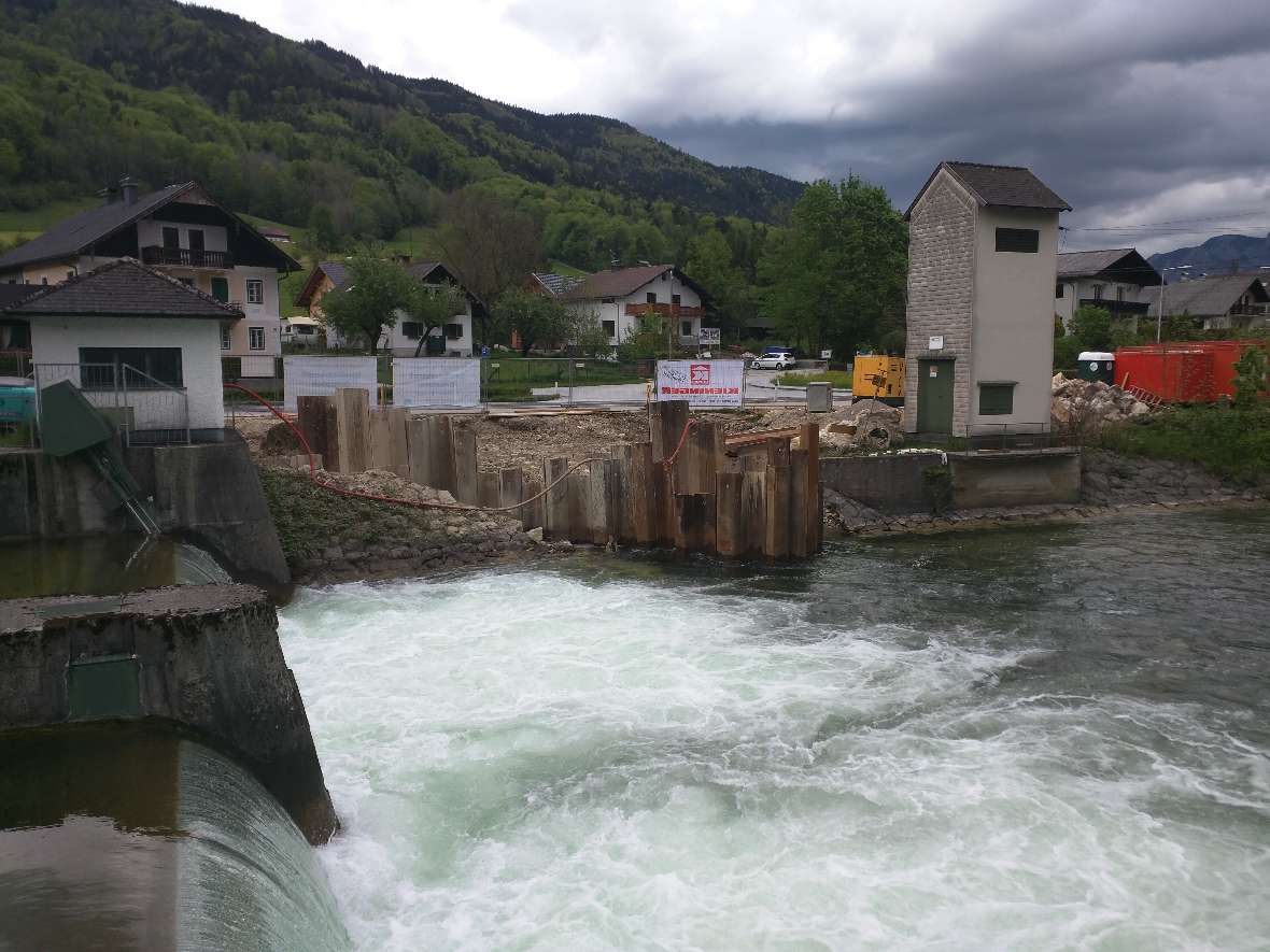 The picture shows the construction site for a fish ladder at a power plant. In the foreground, a small waterfall can be seen, followed by foaming water. In the background are steel sheet pile walls that delimit the construction site. On the right side of the picture there is a transformer house for the power distribution in the adjacent settlement.