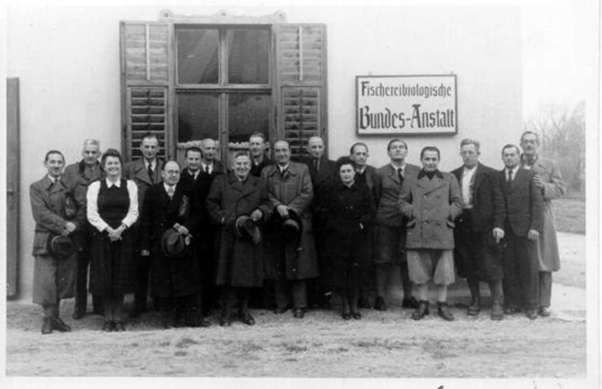 You can see a group photo of the colleagues at the Federal Fisheries Biology Institute in Weissenbach am Attersee. 16 colleagues and 2 female colleagues in front of a window with open shutters, as was typical in the past. On the right is a large sign with the inscription "Fischereibiologische Bundes Anstalt".