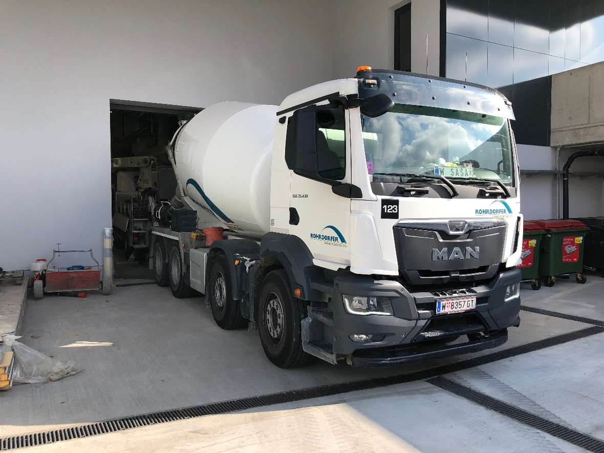 A concrete mixer truck (truck with four axles) is parking in front of the entrance to the hall.