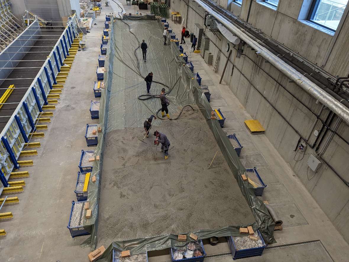 The picture shows the area to be concreted, which is lined with a plastic sheet. Around the area are many blue boxes filled with model stones that hold the formwork. The concrete flows into the formwork via a hose. In the model, five people are busy spreading the concrete and producing the plate.
