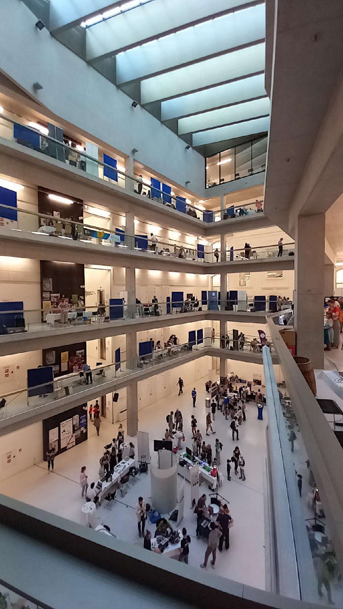 You can see the event space consisting of five levels. An inner courtyard is covered with a glass roof. Corridors run around the inner courtyard. Information booths from many scientific institutions are set up in these corridors. Numerous visitors can be seen around the booths.