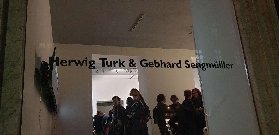 The picture consists of 2 parts. In both parts you can see from the street through the shop windows into the gallery. In the left picture you see many people at the exhibition opening and the names of the artists Herwig Turk and Gebhard Sengmüller written on the glass. On the right picture, the name of the exhibition is written on the glass of the shop window. Directly behind the window is a work of art (a tall man with a long beard made of yellow foam).