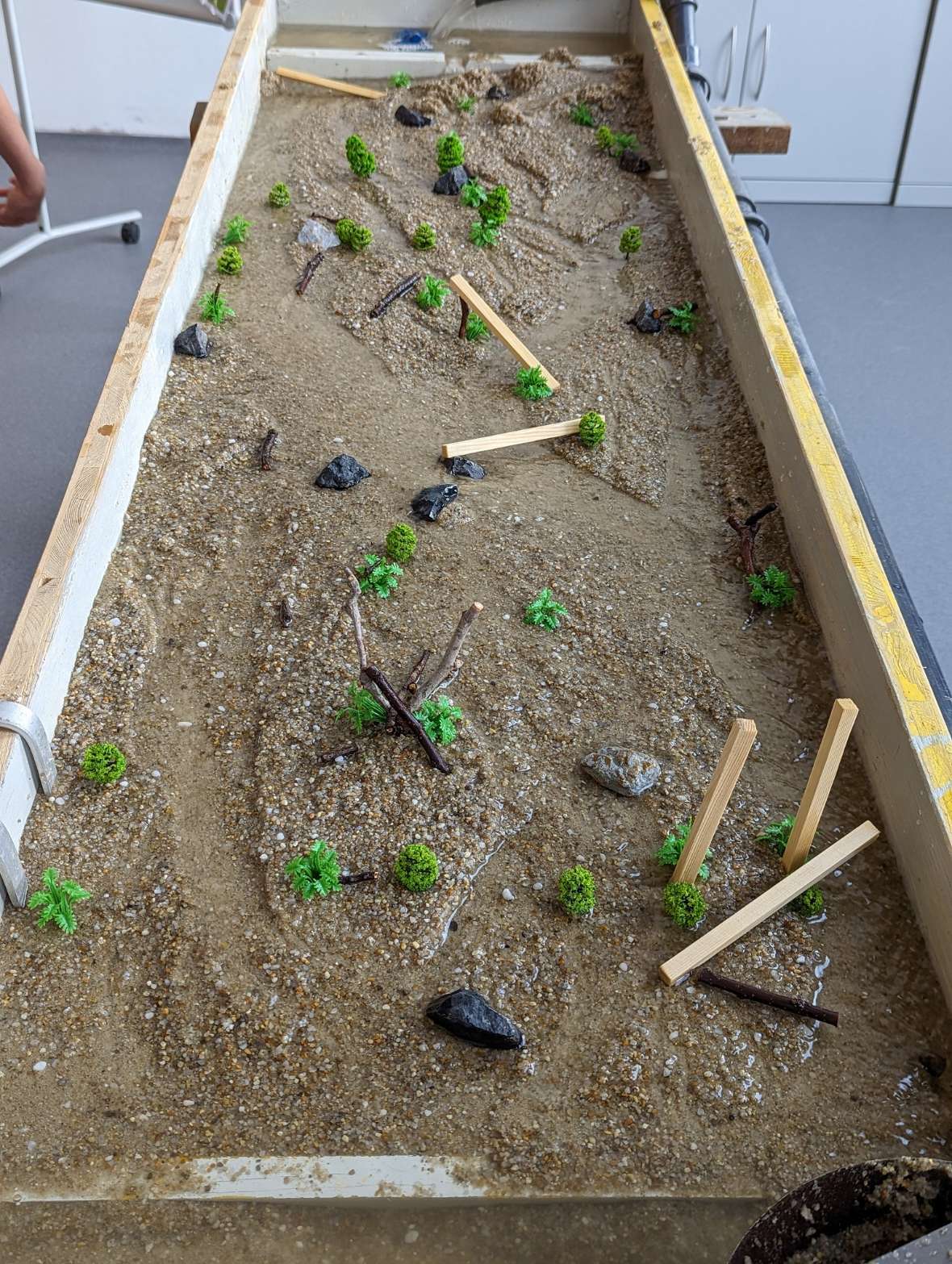 An approximately sixty by two hundred centimetre wooden box is set up inclined and filled with sand. Water runs into the top and drains down through a natural channel in the sand. The water flow can be influenced with coloured stones and small wooden sticks. Six different examples of design can be seen on the photos. Natural, branching courses can be seen, as well as straightening and dams. On some of the examples, children can be seen during the channel design.