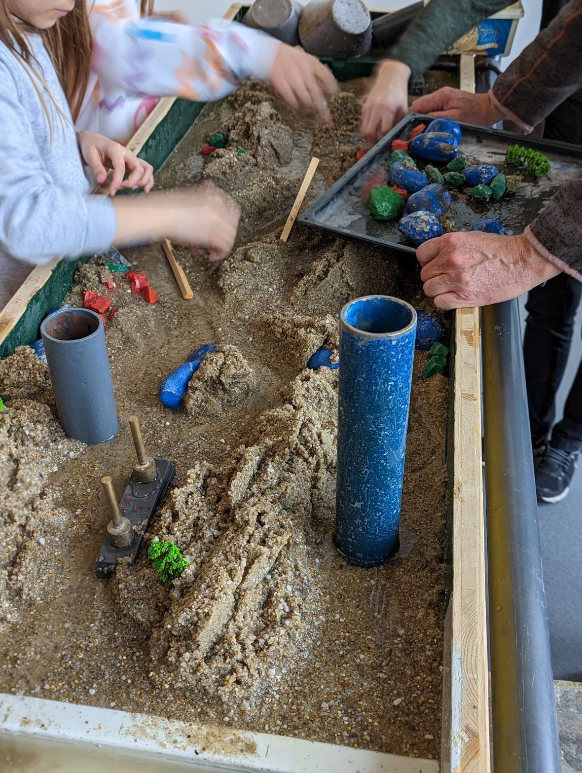 An approximately sixty by two hundred centimetre wooden box is set up inclined and filled with sand. Water runs into the top and drains down through a natural channel in the sand. The water flow can be influenced with coloured stones and small wooden sticks. Six different examples of design can be seen on the photos. Natural, branching courses can be seen, as well as straightening and dams. On some of the examples, children can be seen during the channel design.