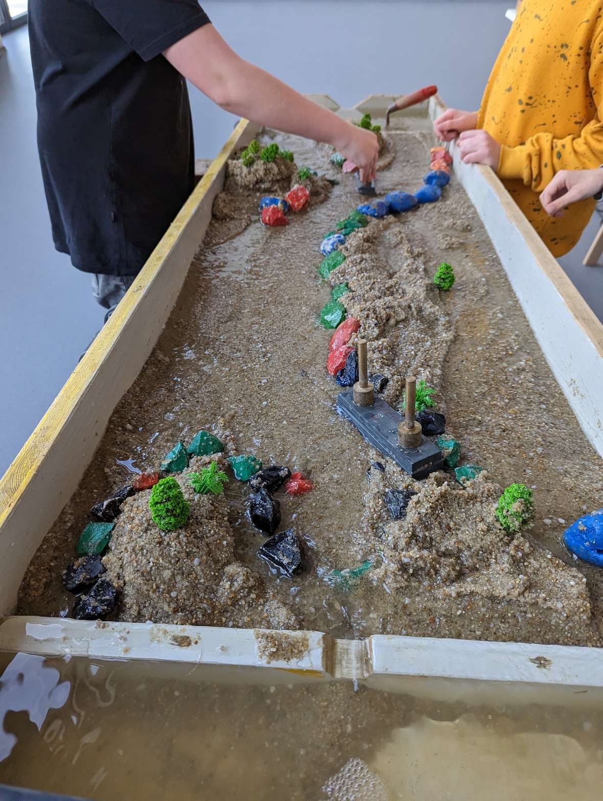 An approximately sixty by two hundred centimetre wooden box is set up inclined and filled with sand. Water runs into the top and drains down through a natural channel in the sand. The water flow can be influenced with coloured stones and small wooden sticks. Six different examples of design can be seen on the photos. Natural, branching courses can be seen, as well as straightening and dams. On some of the examples, children can be seen during the channel design.