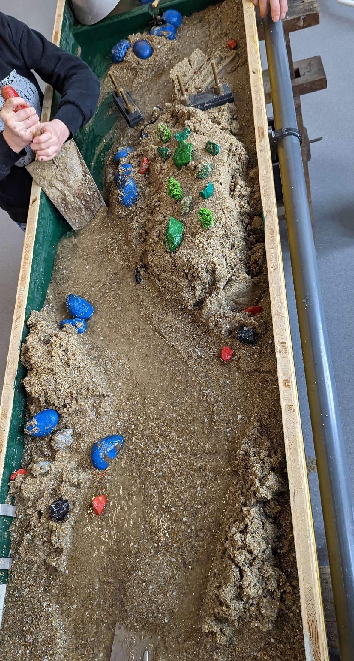 An approximately sixty by two hundred centimetre wooden box is set up inclined and filled with sand. Water runs into the top and drains down through a natural channel in the sand. The water flow can be influenced with coloured stones and small wooden sticks. Six different examples of design can be seen on the photos. Natural, branching courses can be seen, as well as straightening and dams. On some of the examples, children can be seen during the channel design.