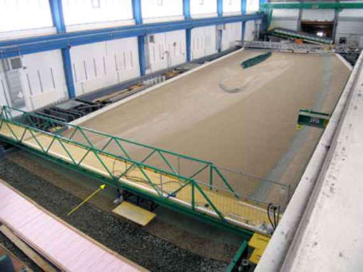 The picture shows the full model seen from above, without water. You can see a large sandy area, which represents the Danube in the model. Across the model a bridge can be seen, which serves for the observation of the experiment.