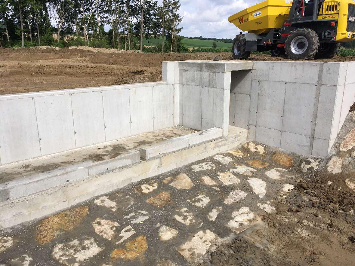 Outlet structure of the research basin. In the front you can see large stones laid in concrete. At the top right is a construction vehicle, further back forest.