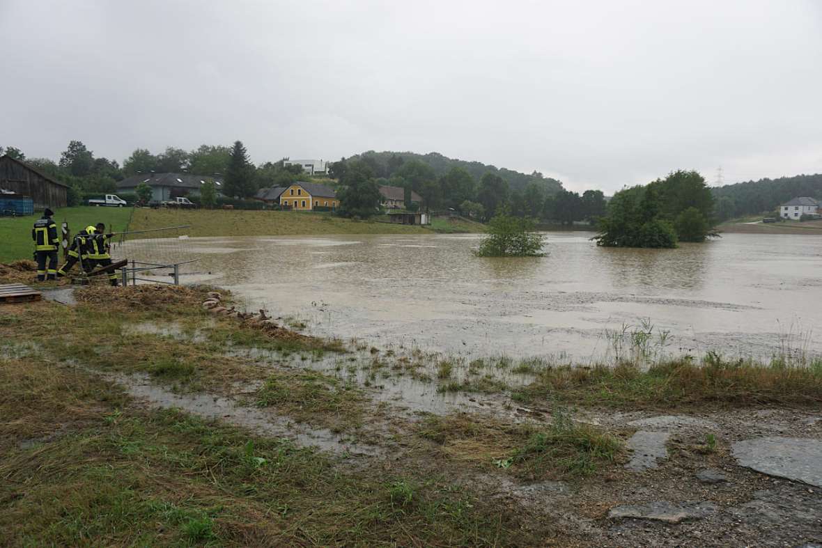 In the foreground you can see a pond filled to the brim. It is the retention basin in Petzenkirchen. It is bad weather, in the background you can still see a few houses. At the left edge of the picture you can see three people in firefighters' clothing working on the drainage of the retention basin.