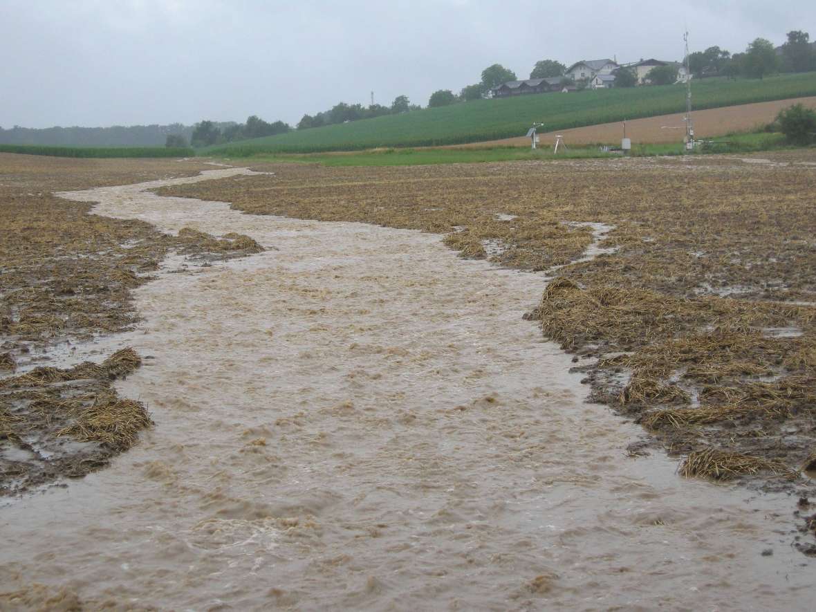 In the picture you can see a hilly landscape in bad weather. On the field in the middle of the picture, due to heavy rainfall, a wide stream has formed and is running downhill, taking a lot of field soil with it.