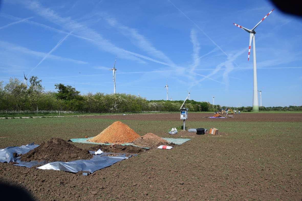 Photo of the establishment of the measuring sites. On a field you can see tarpaulins with earth and new equipment, behind it a wind turbine. On the left is forest in almost cloudless weather.