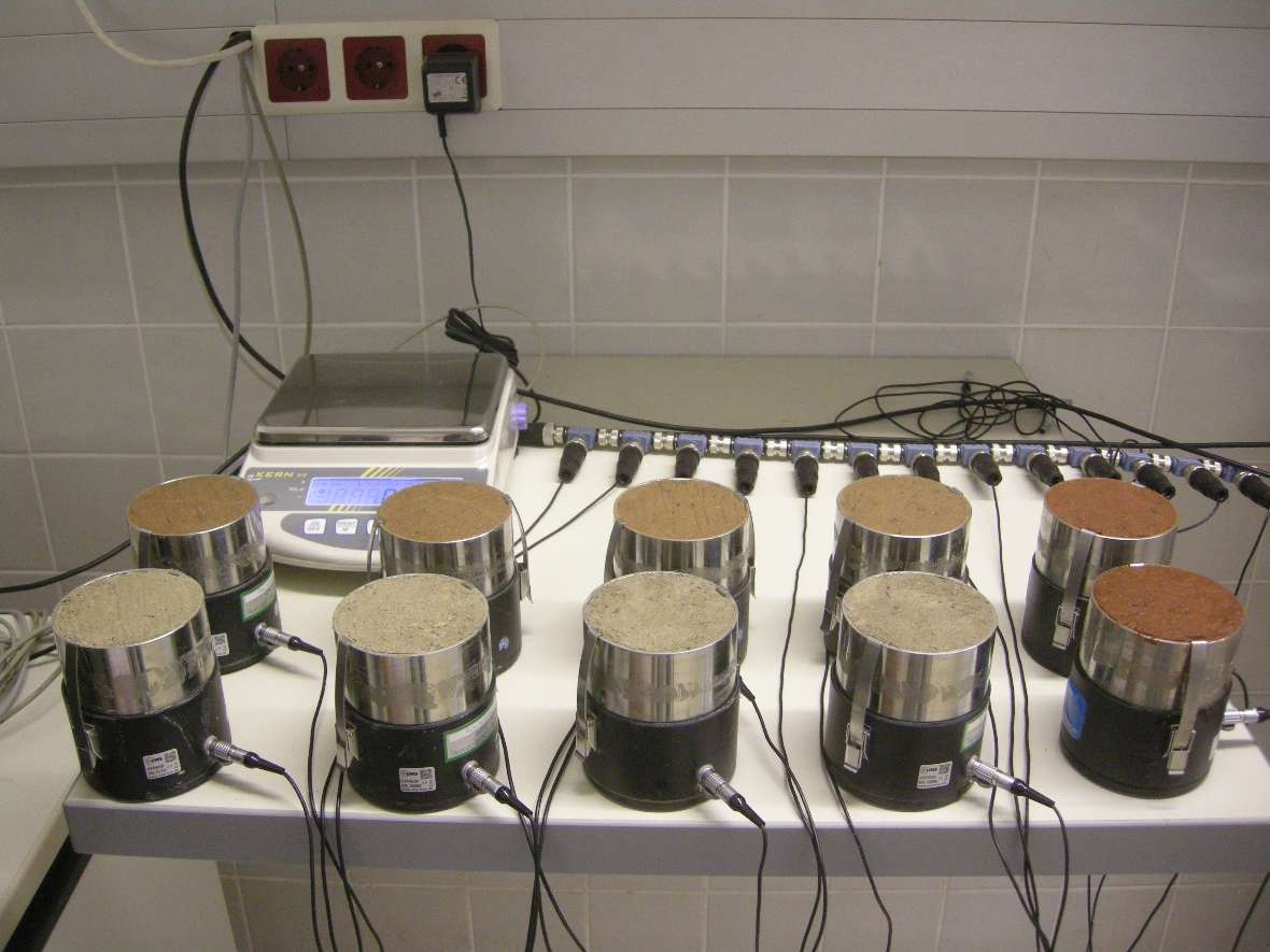In this photo you can see a table with 10 so-called hyprop cylinders arranged in two rows. These cylinders are used to determine the conductivity of soils that are not completely saturated with water. Each cylinder consists of a black lower part and a silver upper part, which contains the soil sample. A cable with a measuring device for conductivity is connected to each cylinder. Behind it is a balance on the table, which is used to regularly measure the change in weight of the cylinders.