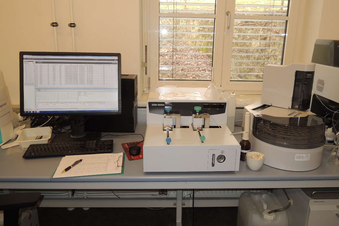 			On a table on the left is a computer with monitor and keyboard, and in the middle and on the right are two large white devices that are used to determine the organic carbon content in a soil sample. For this purpose, the soil sample is burned at a very high temperature in the right-hand device and the carbon content is measured in the device in the middle. The result is the so-called TOC value (acronym for total organic carbon).		