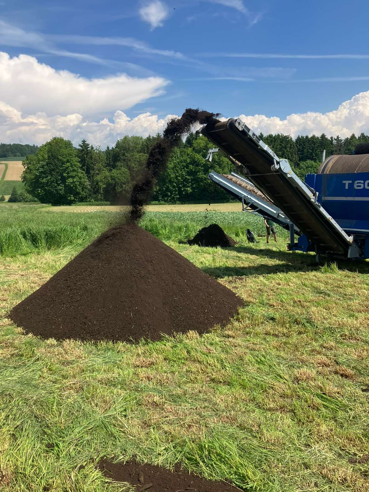 Eine Maschine kippt Erde zu einem Haufen auf eine Wiese, im Hintergrund Bäume und blauer Himmel mit Wolken.