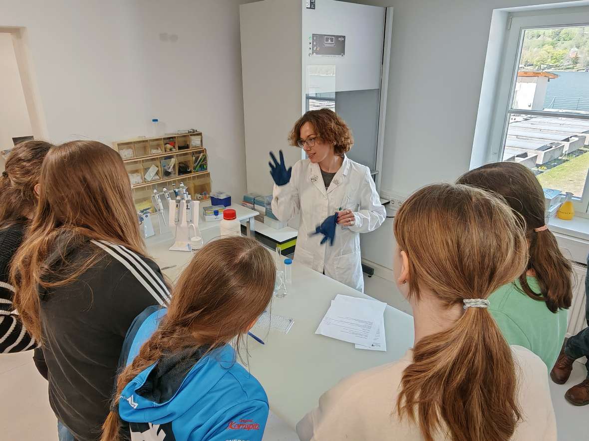 Eine Person im weißen Laborkittel und mit blauen Handschuhen steht an einem Labortisch und spricht zu den Mädchen. Auf dem Tisch liegen Laborutensilien wie Messzylinder, Bechergläser, Pipetten und ein Blatt Papier. 