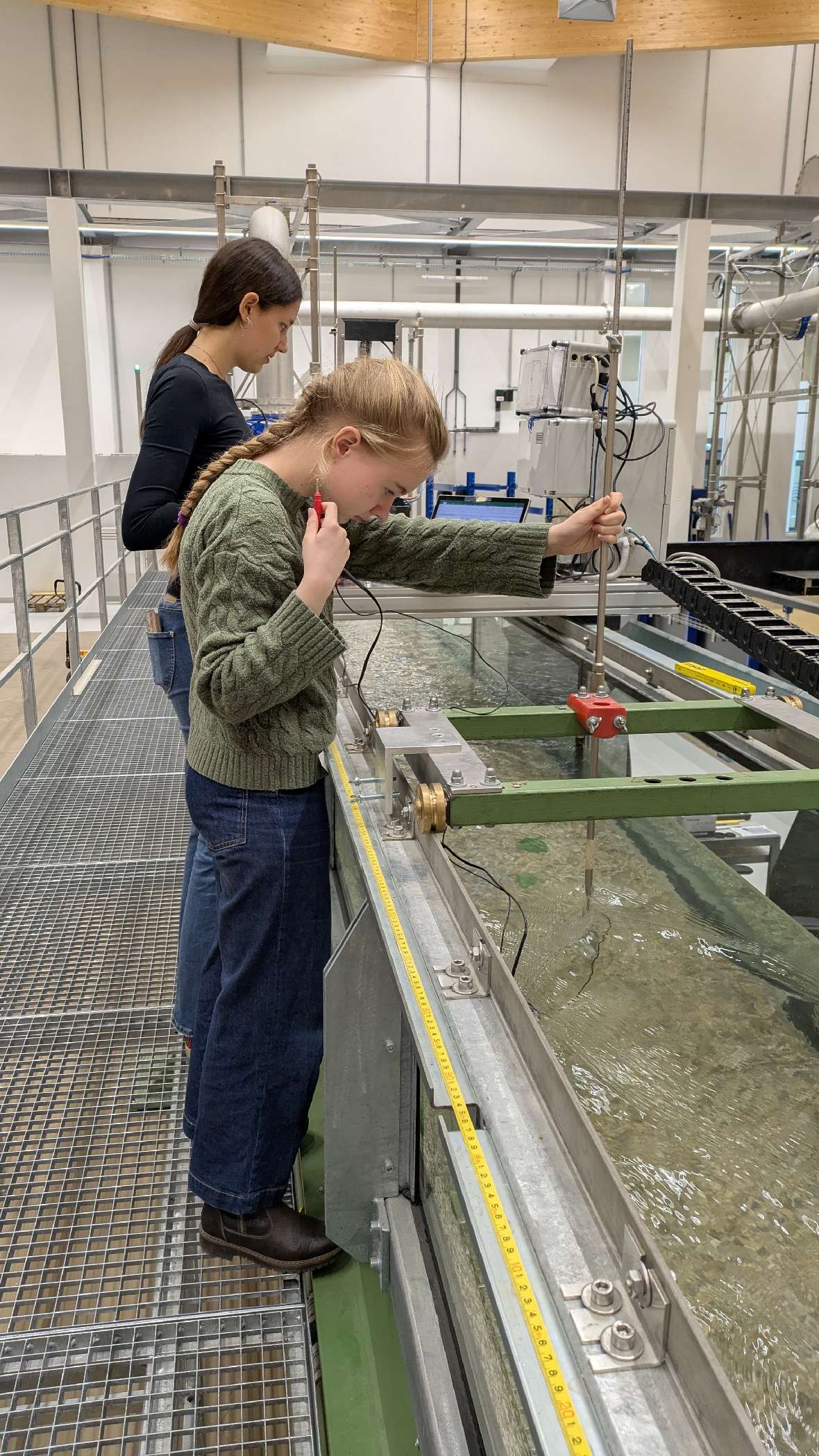 IWB: Zwei Mädchen stehen an einer großen Versuchsrinne, ein Mädchen hält ein Messgerät ins Wasser.