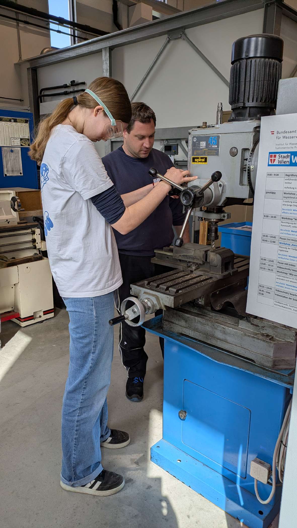 Ein Mädchen und ein BAW-Mitabeiter stehen an einer Werkzeugmaschine in einer Werkstatt, beide tragen Schutzbrillen.