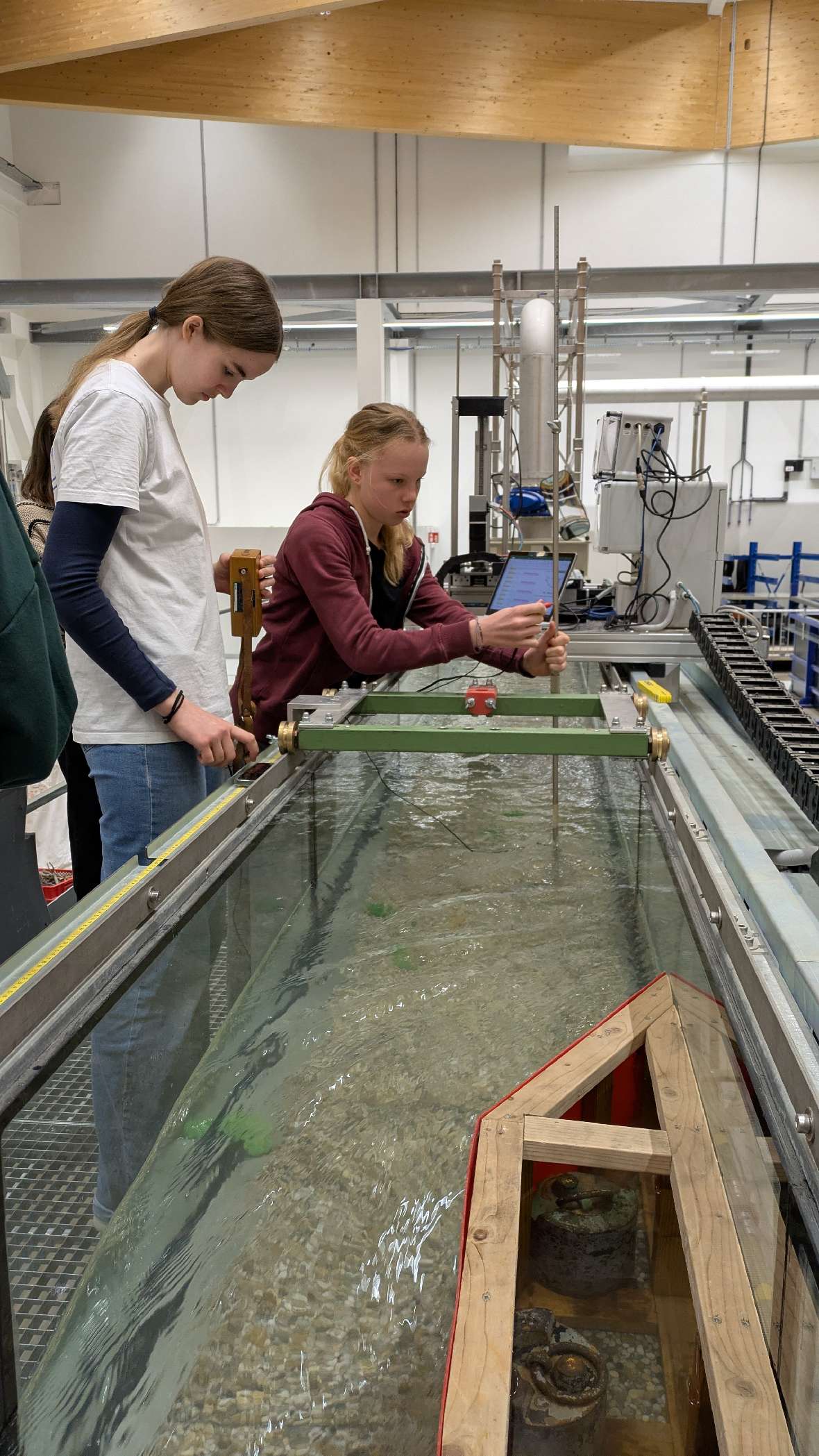 Zwei Mädchen stehen bei einer langen Wasserversuchsrinne mit einem hölzernen Modell im Wasser, im Hintergrund technische Geräte und eine Werkstattumgebung.