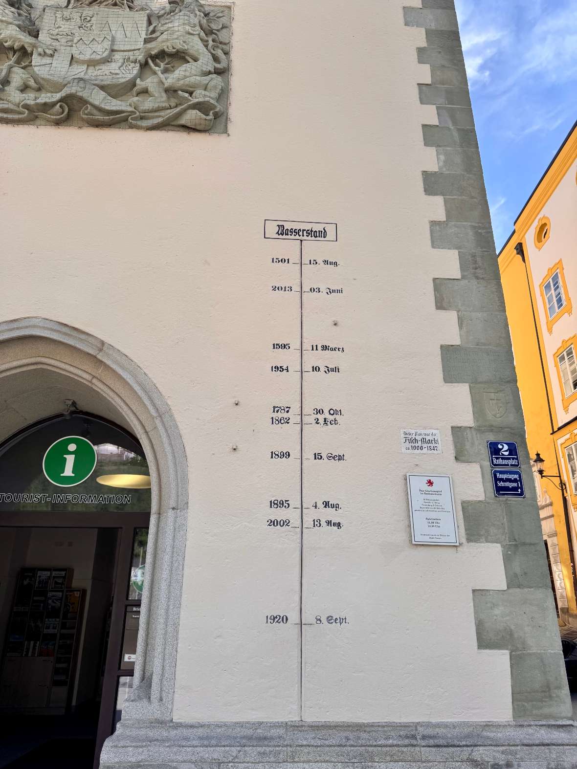 Passau: Wasserstandsanzeige an einer hellen Hauswand mit mehreren historischen Daten und einem Eingang mit grünem Informationssymbol.